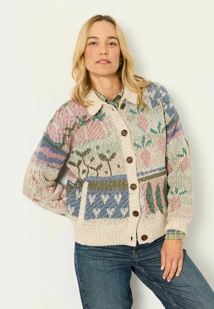 Frau mit blondem Haar, die eine mehrfarbig gemusterte Strickjacke mit braunen Knöpfen und blaue Jeans trägt, steht vor einem schlichten hellen Hintergrund.
