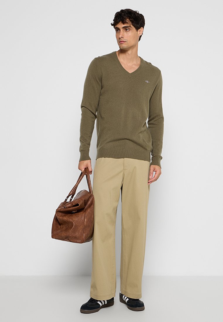 Pull en V en tricot vert olive, pantalon large beige, sac de voyage en cuir marron, et baskets noires avec des bandes blanches.