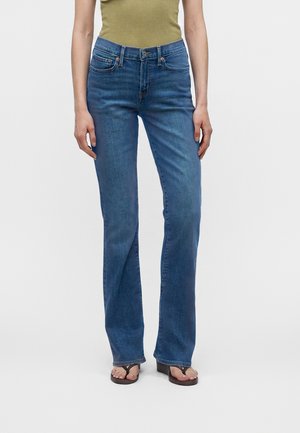 Γυναίκα που φοράει ψηλόμεσα μπλε τζιν bootcut, ελιάς τοπάκι και μαύρα σανδάλια με τακούνι, στέκεται μπροστά σε απλό ανοιχτό φόντο.