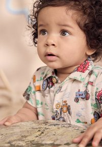 Chemise à col en tissu crème avec des motifs colorés de tracteurs et d'animaux de ferme. Comprend des boutons et des manches courtes, surface texturée visible.