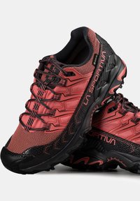 Červené a černé trailové běžecké boty s prodyšným svrškem, texturovanými akcenty a robustní podrážkou. Obsahují vyztuženou špičku a elastické tkaničky.
