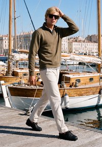 Homme blond avec des lunettes de soleil, un pull olive, un pantalon blanc et des chaussures noires marchant sur un quai en bois à côté de voiliers en bois amarrés dans une marina.