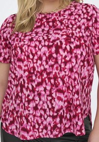 Kobieta ubrana w krótki rękaw różową i czerwoną bluzkę w abstrakcyjny wzór, z naszyjnikiem z złotego łańcuszka, w zestawie z ciemnymi spodniami.