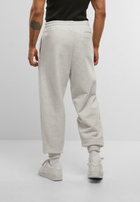 Osoba v svetlosivých teplákoch, bielych teniskách a bielych ponožkách, stojaca proti jednoduchému svetlosivému pozadiu, pohľad zozadu.