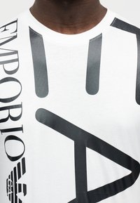 Λευκό βαμβακερό T-shirt με μεγάλα μαύρα γραφικά σε σχήμα χεριού και έντονο κείμεν ο, με στρογγυλή λαιμόκοψη και απαλό υλικό.