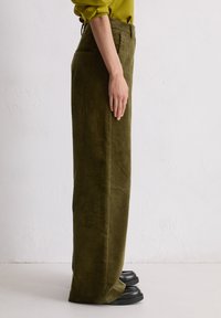 Olijfgroene corduroy broek met een rechte pasvorm, voorzien van verticale ribbels en zijzakken. Draagt met zwarte platform schoenen.