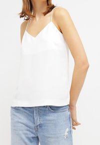 Camicetta bianca con scollatura a V, spalline sottili regolabili e finitura satinata liscia. Abbinata a jeans azzurri chiari strappati.