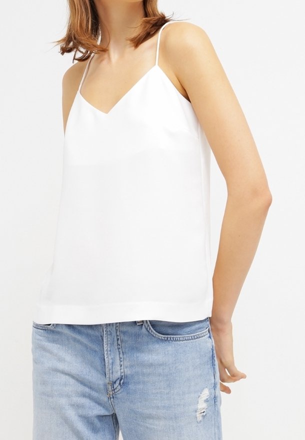 Camicetta bianca con scollatura a V, spalline sottili regolabili e finitura satinata liscia. Abbinata a jeans azzurri chiari strappati.