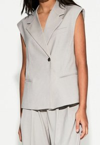 Ljust grå ärmlös blazer med notch-krage, enkel knappstängning, två framfickor och en mjuk, slät yta. Slät tygfinish.