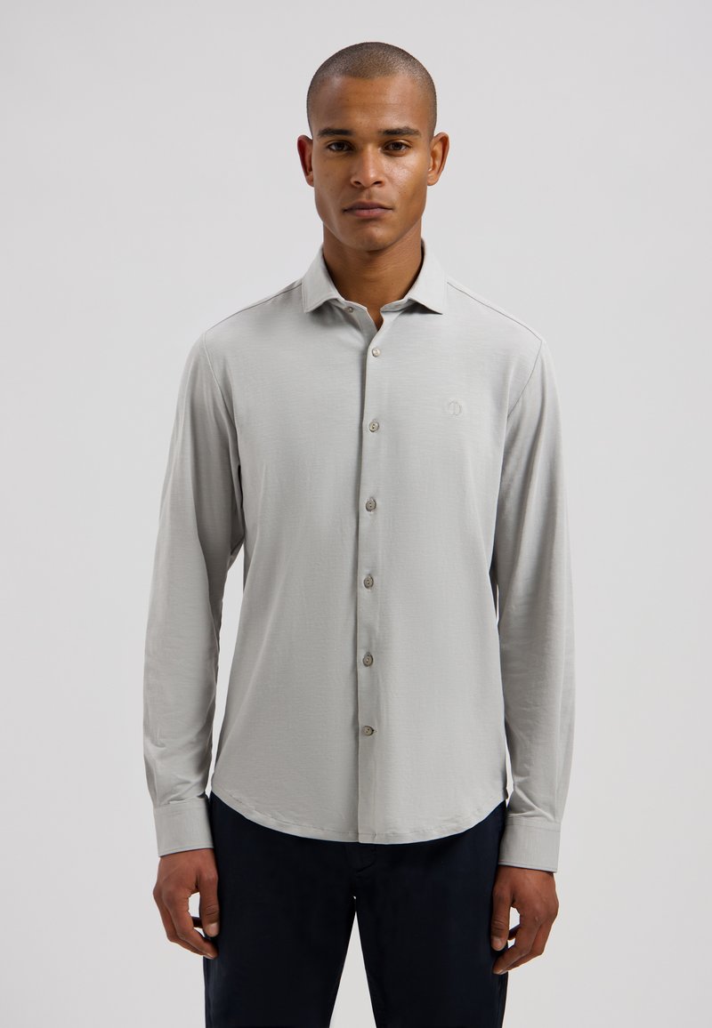 Homme aux cheveux courts portant une chemise à manches longues gris clair boutonnée et un pantalon foncé, debout devant un fond clair uni.