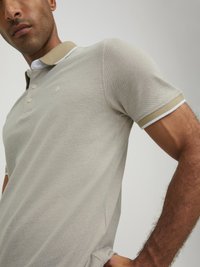 Hellbeige Polohemd aus strukturiertem Stoff mit einem kontrastierenden braunen Kragen und weißen Akzenten an den Ärmeln und dem Kragen.