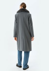Abrigo de lana gris con cuello de piel, diseño recto, corte a media longitud y bolsillos laterales, combinado con vaqueros de color azul claro y zapatos negros.