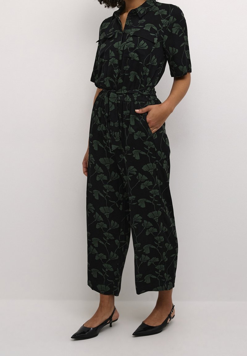 Combinaison noire avec un motif de feuilles vertes, manches courtes, deux poches poitrine, taille froncée et pantalons à jambes larges. Associée à des escarpins noirs à bride arrière.