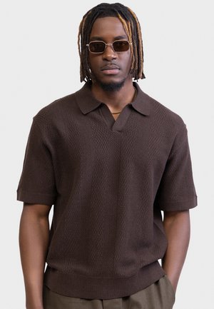 Mand med korte dreadlocks og solbriller iført en mørkebrun tekstureret kortærmet strikket polo-shirt og olivengrønne bukser.