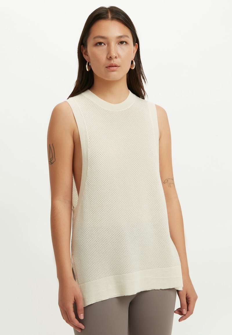 Kvinna med långt mörkt hår bär en ärmlös, off-white stickad topp med öppna sidor, stora ringörhängen och taupefärgade leggings mot vit bakgrund.