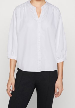 Personne portant un chemisier blanc à manches longues boutonné avec un col en V, associé à un pantalon noir, debout devant un fond uni.