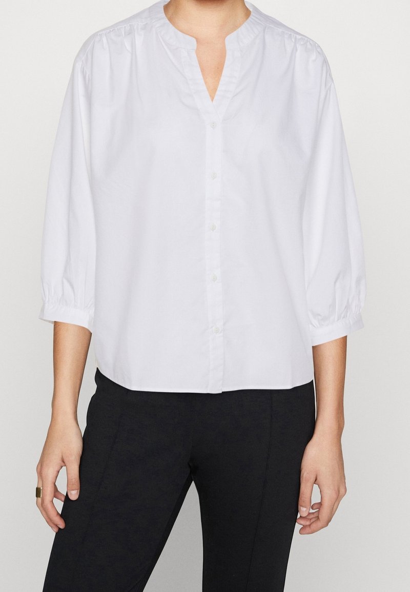 Personne portant un chemisier blanc à manches longues boutonné avec un col en V, associé à un pantalon noir, debout devant un fond uni.
