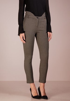 Kobieta ubrana w brązowe, dopasowane, teksturowane spodnie do kostek, czarną przejrzystą bluzkę z długim rękawem oraz czarne buty na wysokim obcasie, stojąca na beżowej podłodze.