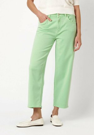 Person wearing light green straight-leg pants and white loafers, with one hand in pocket, standing against a plain background.