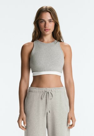 Femme aux longs cheveux bruns portant un crop-top gris sans manches et un pantalon de survêtement gris avec un cordon, debout devant un fond uni.