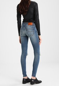 Kobieta ubrana w niebieskie skinny jeans z niewielkimi przetarciami oraz czarną bluzkę z długim rękawem, stojąca tyłem do aparatu na białym tle.