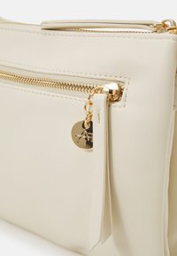 Skórzana torba w kremowym kolorze z złotym zamkiem błyskawicznym, wyposażona w odpinany brelok i elegancki design. Teksturowana powierzchnia z minimalnymi zdobieniami.