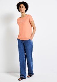 Meruňkový tričko s krátkým rukávem s potiskem "NEKONEČNÝ SEN" v světlejším písmu. V kombinaci s širokými džínovými kalhotami modré barvy a černými pantoflemi.