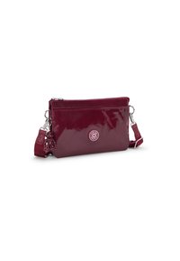 Sac à bandoulière en verni bordeaux avec une forme arrondie, fermeture éclair, et patch logo. Comprend une bandoulière détachable et un accent charm.