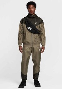 Kurtka przeciwwiatrowa Nike i spodnie dresowe w oliwkowej zieleni z czarnymi akcentami; posiada zamek z przodu, elastyczne mankiety oraz szczegóły z logo. Czarna torba na ramię.