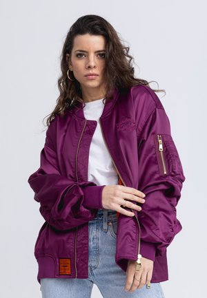 Purple bomber jacket with a glossy satin finish, ribbed cuffs, and side pockets. Features a zippered pocket on the sleeve and an orange lining.