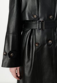 Black leather coat featuring a double-breasted design with brown buttons, a belted waist, and a smooth texture.