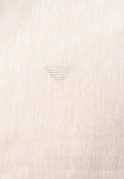 Υφασμα λινό σε ανοιχτό μπεζ χρώμα με μια διακριτική ανάγλυφη γεωμετρική λογότυπο στο κέντρο του κάδρου.