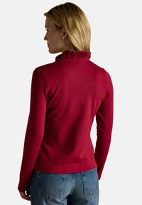 Frau mit blonden Haaren, die ein rotes Langarmoberteil mit gerüschtem Kragen und blaue Jeans trägt, mit dem Rücken zu einer schlichten Hintergrund.