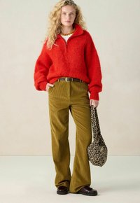 Blonde Frau in einem roten flauschigen Zip-up-Pullover, olivgrünen Cordhosen, dunklem Gürtel, braunen Schuhen, mit einem bernsteinfarbenen Halsband, die eine Tasche mit Leopardenmuster hält.