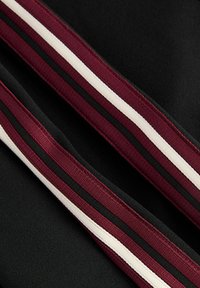 Crni materijal s dva dijagonalna prugasta naglaska u bordo i crnoj boji uz bijelu prugu; glatke teksture i sportski dizajn.
