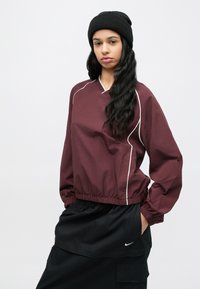 Bordový top s dlhým rukávom a výstrihom do V, so svetlými bielymi lemami a elastickým lemom, doplnený čiernou čiapkou a tmavými nákladnými nohavicami.