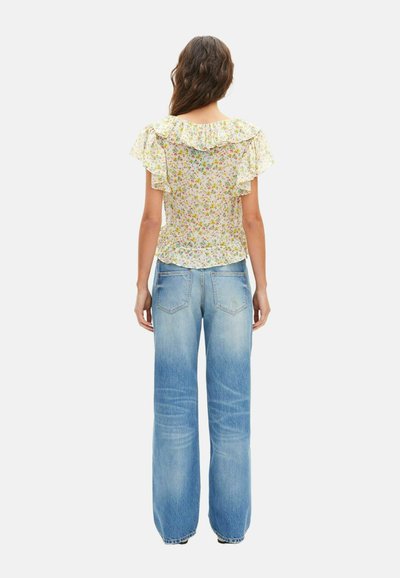 Mujer vista de espaldas con una blusa floral con volantes de manga corta y vaqueros de pierna ancha en azul claro sobre un fondo blanco.