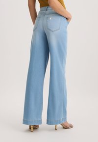 Pantaloni wide-leg in denim blu chiaro con vita alta, dotati di due tasche posteriori e un orlo grezzo, indossati con sandali con tacco.