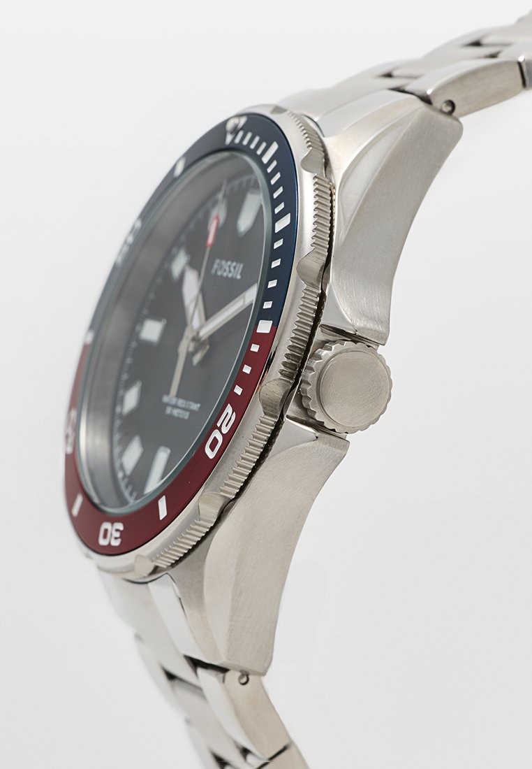 Reloj Fossil con caja de acero inoxidable, esfera negra, bisel rojo y azul, y pulsera de metal plateado. Presenta marcadores de hora prominentes y corona.