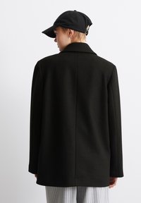 Černý vlněný kabát s klopovým límcem, přímým střihem a dlouhými rukávy, doplněný černou čepicí. Má hladkou texturu a minimální detaily.