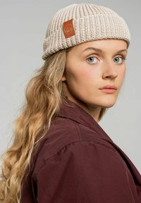 Beżowa dziana czapka z pr�ążkowanym wzorem i skórzaną naszywką z logo górskim, noszona przez osobę z długimi falowanymi włosami i burgundową kurtką.