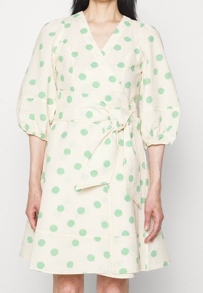 Femme portant une robe portefeuille crème à pois verts, manches trois-quarts bouffantes et nœud à la taille.