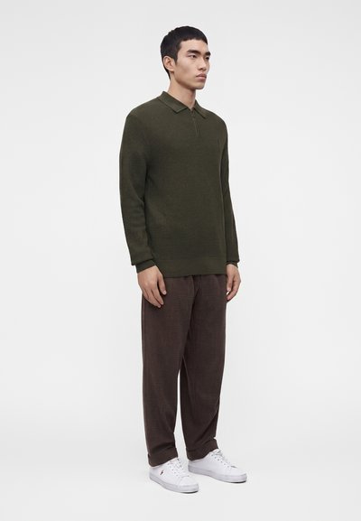 Jeune homme debout, portant un pull en maille vert olive, un pantalon ample marron à carreaux et des baskets blanches sur un fond clair uni.
