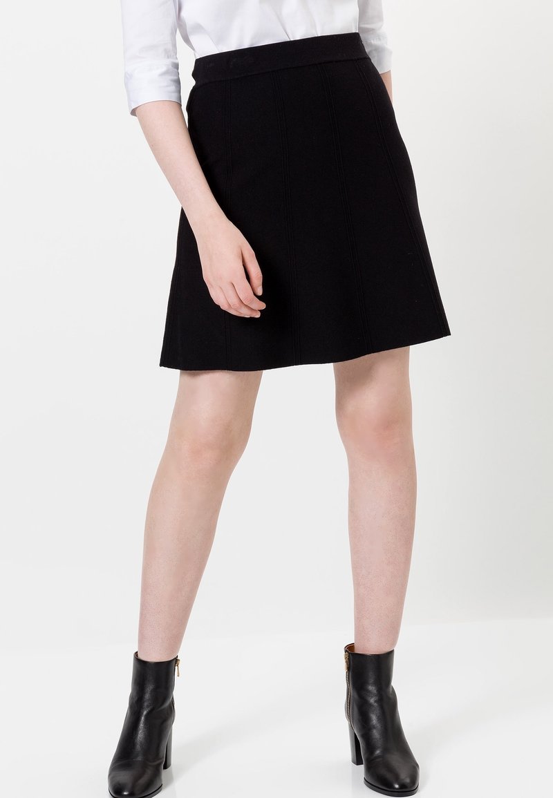 Personne portant une jupe noire évasée, une chemise blanche à manches longues, et des bottines noires à talons hauts, debout devant un fond blanc.
