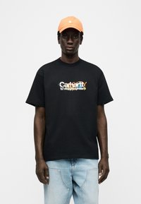 Νεαρός άνδρας με πορτοκαλί καπέλο, μαύρο μπλουζάκι με πολύχρωμο κείμενο και ανοιχτό μπλε τζιν, στέκεται μπροστά σε λευκό φόντο.