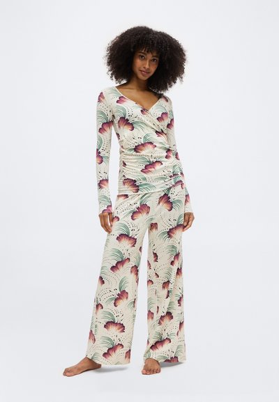 Γυναίκα με σγουρά μαλλιά που φοράει σετ ρούχων με κρεμ χρώμα και λουλουδάτο σχέδιο, κορυφή τύπου wrap και παντελόνα με φαρδιά μπατζάκια, όρθια ξυπόλητη μπροστά σε λευκό φόντο.