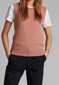 Ärmlös rosa stickad väst med texturmönster, bärs över en vit T-shirt. Botten har en ribbad design och fickor på sidorna.