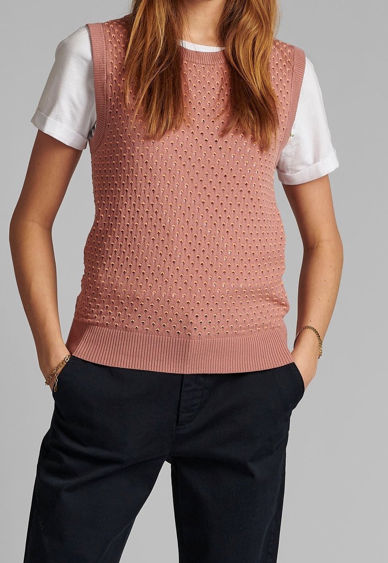 Ärmlös rosa stickad väst med texturmönster, bärs över en vit T-shirt. Botten har en ribbad design och fickor på sidorna.