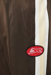 Bruine sportbroek met een witte streep. Heeft een rode geborduurde patch van een springende vos. Glad, lichtgewicht materiaal.