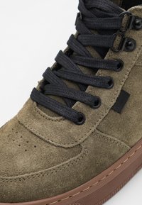 Zielone zamszowe trampki z czarnymi sznurowadłami, okrągłym noskiem, subtelnymi perforacjami i gumową podeszwą z brązowym akcentem na krawędzi.